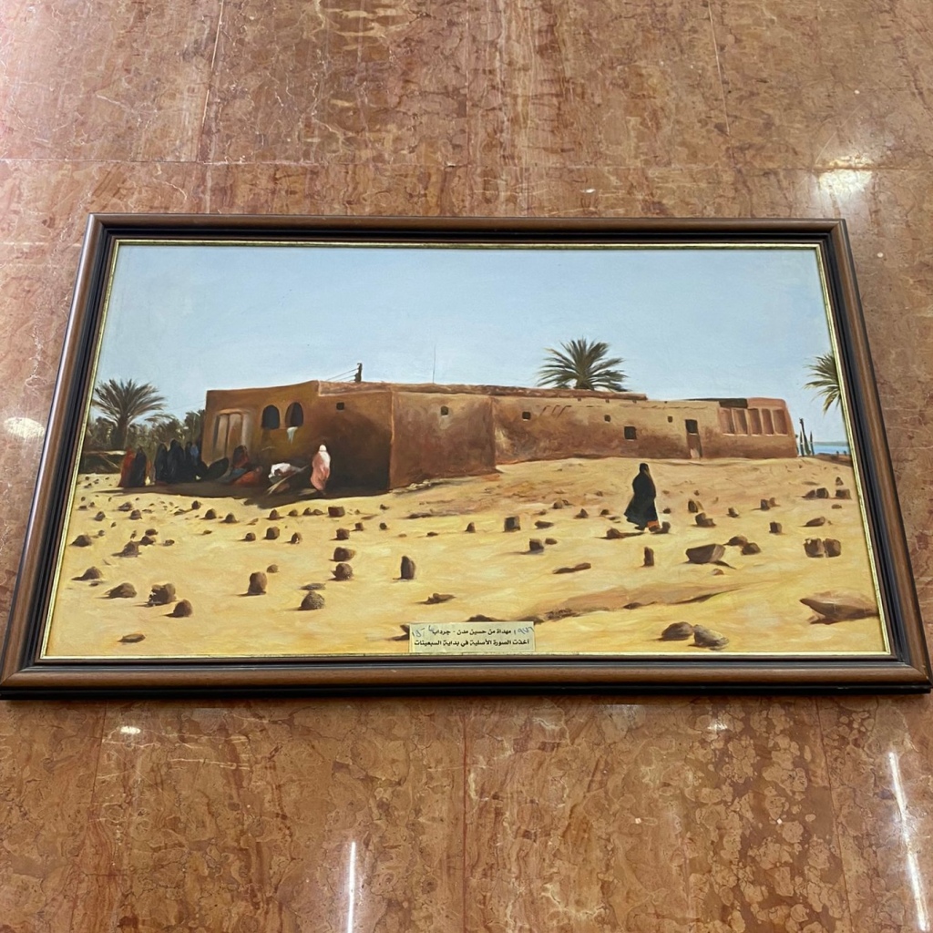 A painting of the old shrine against a marble wall. The painting shows a mudbrick building in an L-shape and a large graveyard. A woman walks between the graves, which stick out from the sand like jagged teeth. The sea is glimpsed in the bacround.
